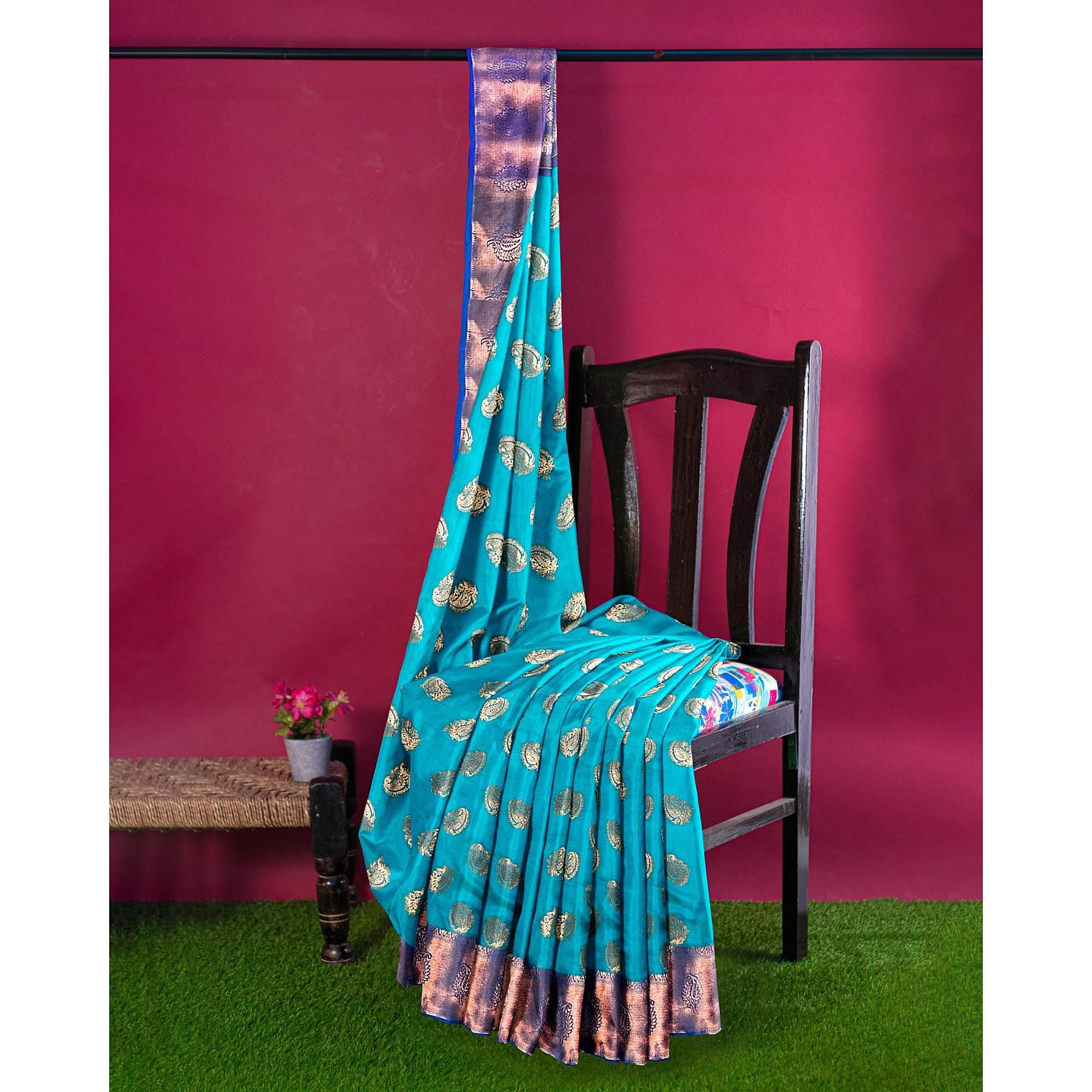 Turquoise handwoven Kanchi silk saree with gold zari work, draped over a chair against a pink backdrop.